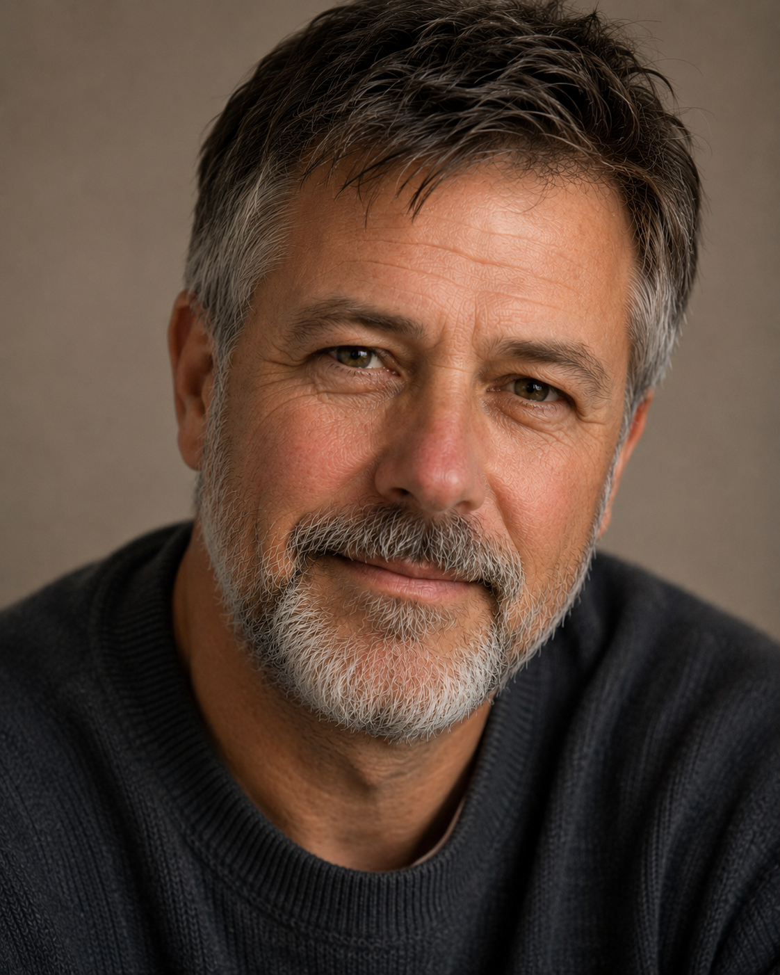 A middle-aged man with short, slightly graying hair and glasses sits at a table, wearing a light-colored button-up shirt. He has a mustache and is smiling gently, with a notepad and pen visible in his shirt pocket.