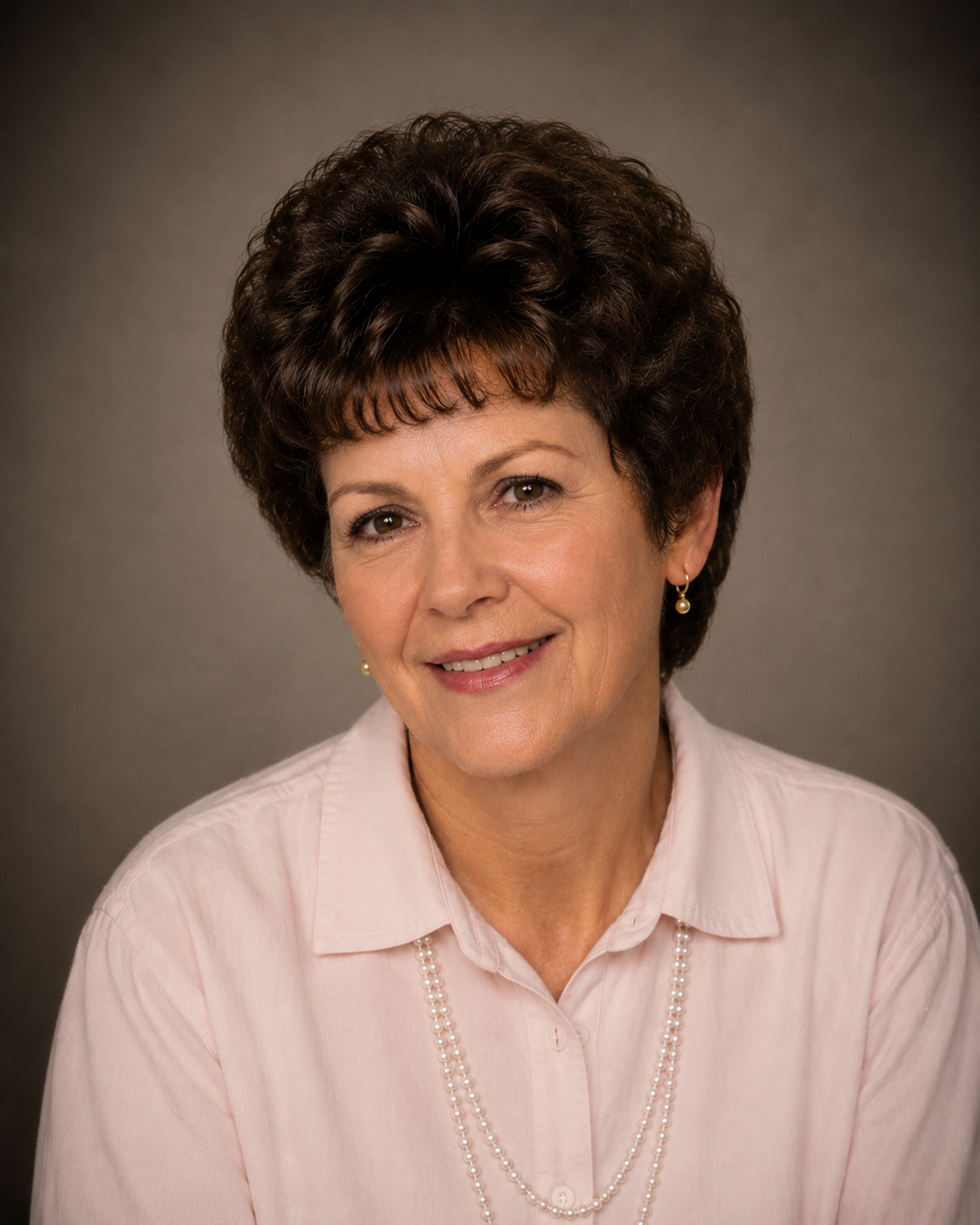 A middle-aged woman with curly brown hair smiles warmly. She is wearing a light pink shirt and a string of pearls, with a simple background.